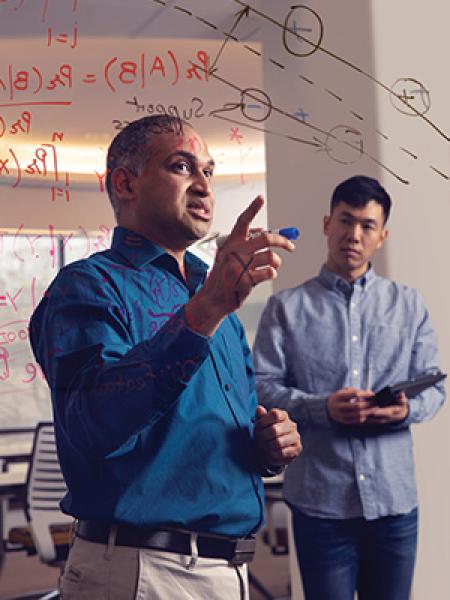 Man gesturing at glass board filled with numbers with a young man standing in the background