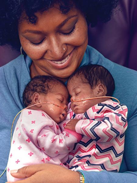 Black woman holding two sleeping babies wearing pink patterned sleepers and with nasal tubes facing each other